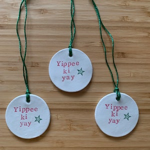 May include: Three white, round ornaments with green string. Each ornament has the phrase "Yippee ki yay" in red text and a green star. The ornaments are displayed on a wooden surface.