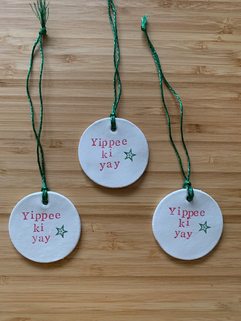 May include: Three white, round ornaments with green string. Each ornament has the phrase "Yippee ki yay" in red text and a green star. The ornaments are displayed on a wooden surface.