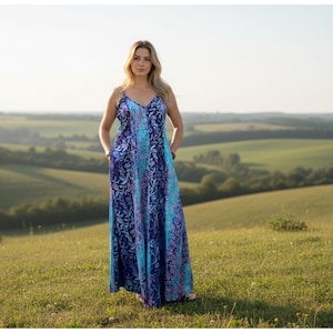 May include: A long, flowing dress with a blue and purple floral pattern. The dress has thin straps and a V-neckline. The person is standing in a field with rolling hills in the background.