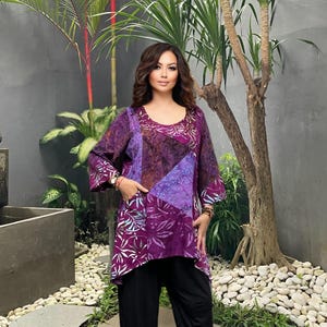 May include: A purple tunic with a patchwork design featuring shades of purple, brown, and white floral patterns. The tunic has long sleeves and a scoop neckline. The model is wearing black trousers. The background includes plants and a water feature.