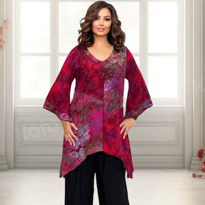 May include: A red and pink floral tunic with a V-neck and flared sleeves. The tunic is worn with black trousers. The garment has an asymmetrical hemline. The model is wearing black trousers.