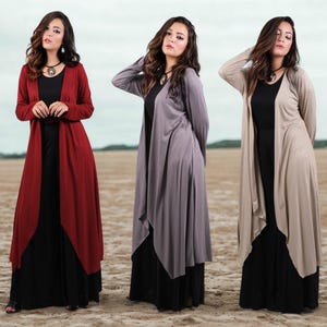 May include: Three long, flowing dresses in a studio setting. Each dress features a black base layer, with a long, open-front cardigan in red, gray, and beige. The models are wearing necklaces and standing on a sandy surface.