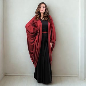 May include: A woman wearing a long, flowing black dress with a matching belt. A draped, burgundy-colored open-front cardigan is worn over the dress. The outfit is accessorized with a necklace. The background is a plain white wall.