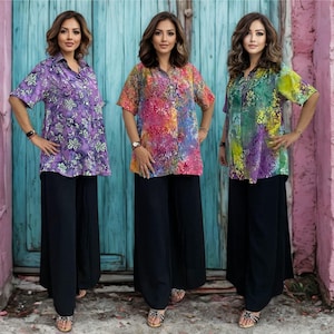 May include: Three women wearing colorful, short-sleeved blouses and black palazzo pants. The blouses feature vibrant patterns in purple, pink, and green. The women are standing in front of a blue door and a pink wall.