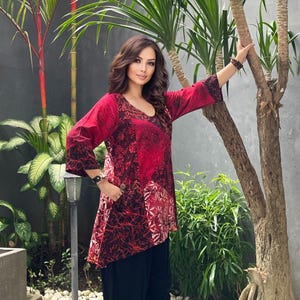 May include: A red and black patterned tunic with three-quarter sleeves and a V-neck. The tunic has an asymmetrical hemline and is paired with black trousers. The model is standing near a tree and tropical plants.