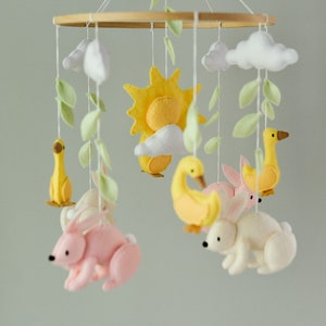 May include: A wooden baby mobile with felt animals and clouds. The mobile features a yellow sun, two yellow ducks, two white bunnies, and a pink bunny. The mobile is hanging from a wooden ring.