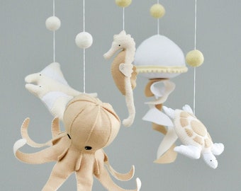 May include: A beige and white felt baby mobile with a seahorse, octopus, turtle, jellyfish, and white pom poms hanging from a wooden ring.