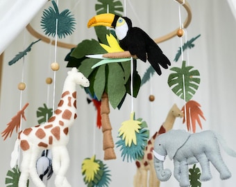 May include: A wooden baby mobile with a jungle theme. It features a toucan, giraffe, elephant, and palm tree, all made of felt. The mobile is suspended from a wooden ring.