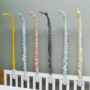 May include: Five colorful fabric-covered crib rail guards in yellow, light blue, pink, gray, and white. The guards are attached to a white crib rail.