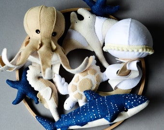 May include: A felt sea life mobile featuring an octopus, stingray, seahorse, turtle, whale shark, starfish, and a jellyfish. The creatures are in shades of beige, white, and navy blue, arranged within a wooden hoop. The jellyfish has a yellow trim.