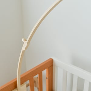May include: A wooden baby crib mobile arm attached to the side of a wooden crib. The arm is curved and has a small knob at the end.
