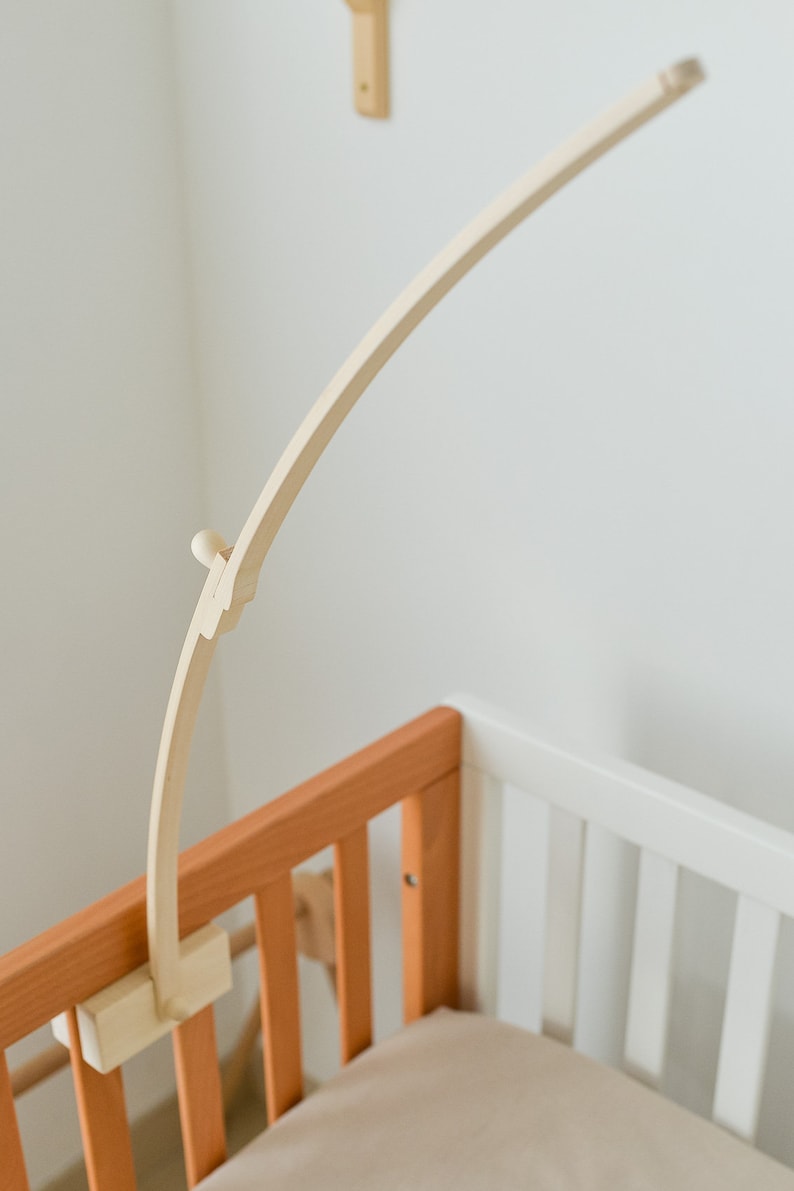 May include: A wooden baby crib mobile arm attached to the side of a wooden crib. The arm is curved and has a small knob at the end.
