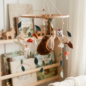 May include: A wooden baby mobile featuring woodland creatures. The mobile includes a white rabbit, a brown bear, a deer, and an owl, along with felt leaves and mushrooms. The mobile is suspended from a wooden ring.