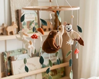 May include: A wooden baby mobile featuring woodland creatures. The mobile includes a white rabbit, a brown bear, a deer, and an owl, along with felt leaves and mushrooms. The mobile is suspended from a wooden ring.