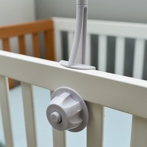 May include: White plastic crib toy attachment with a round knob and a white plastic arm. The attachment is mounted on a white crib rail.