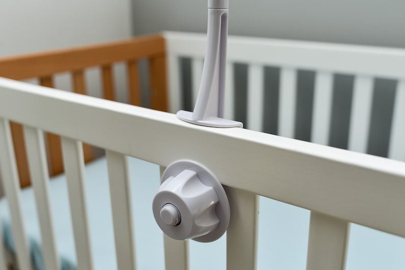 May include: White plastic crib toy attachment with a round knob and a white plastic arm. The attachment is mounted on a white crib rail.
