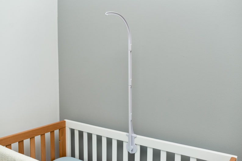 May include: White plastic crib mobile arm with a curved hook at the top. The arm is attached to the side of a crib with a clamp.