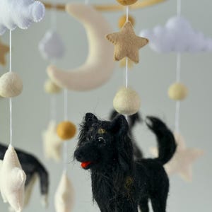May include: A baby mobile with a wooden hoop, featuring a black dog, stars, clouds, and a crescent moon. The mobile includes felted elements in neutral colours. The dog has yellow accents on its legs.