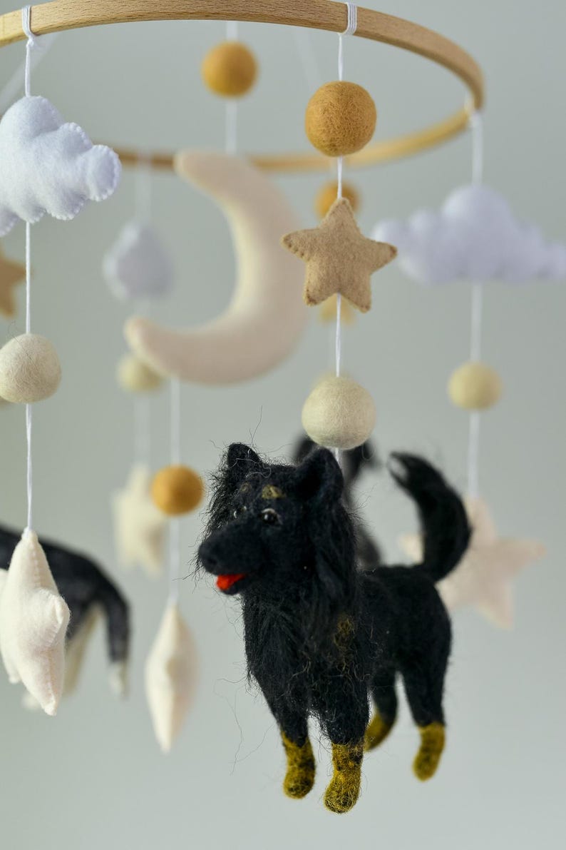 May include: A baby mobile with a wooden hoop, featuring a black dog, stars, clouds, and a crescent moon. The mobile includes felted elements in neutral colours. The dog has yellow accents on its legs.