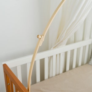 May include: A wooden baby crib mobile arm, made of natural wood, is attached to the side of a white crib. The arm is curved and has a small wooden knob at the top.