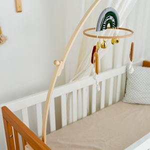 May include: A wooden baby crib mobile with a rainbow, stars, and bells hanging from it. The mobile is attached to a wooden arm that is mounted to the side of the crib. The crib is white with a brown wooden headboard and footboard.