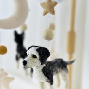 May include: A handcrafted baby mobile with felt dog figures in black, white, and grey. The mobile also features felt stars, moons, clouds, and spherical accents in neutral tones, suspended from a wooden frame.