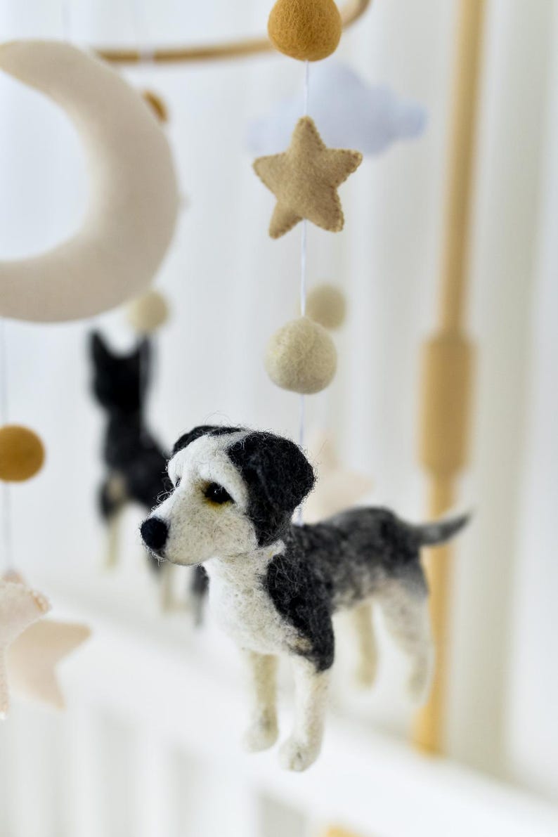 May include: A handcrafted baby mobile with felt dog figures in black, white, and grey. The mobile also features felt stars, moons, clouds, and spherical accents in neutral tones, suspended from a wooden frame.