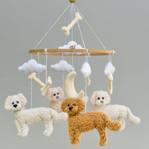May include: A nursery mobile with cream and brown toy dogs, clouds, a crescent moon, and bone shapes. The mobile is suspended from a wooden hoop. The soft toys hang from white strings, creating a whimsical design.