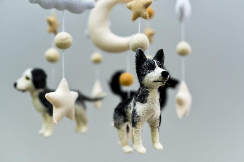 May include: A baby mobile featuring felted dog figures in black and white, along with stars, moons, and clouds. The nursery decor is suspended by white strings, creating a whimsical and gentle atmosphere.