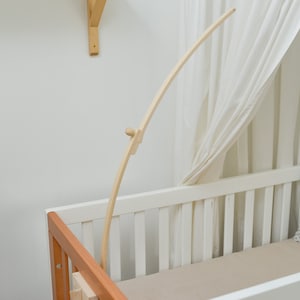 May include: A wooden baby crib mobile arm attached to a white crib with a beige fitted sheet and a gray patterned pillow. The arm is curved and extends over the crib.
