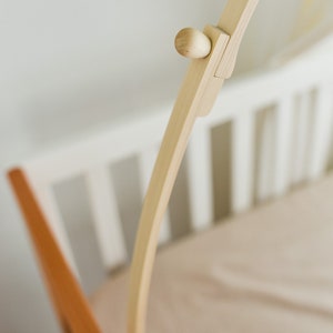 May include: A wooden crib mobile stand with a natural finish. The stand is made of two curved pieces of wood that meet at the top. The stand is designed to be attached to the side of a crib.