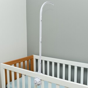 May include: White baby crib mobile arm with a curved arm and a white plastic base. The arm is attached to the side of a white crib with a wooden top rail.