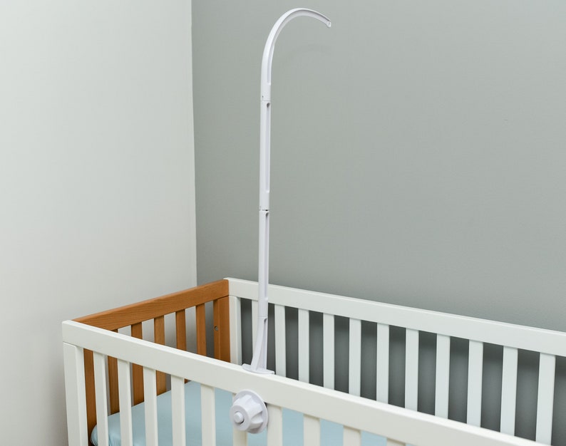 May include: White baby crib mobile arm with a curved arm and a white plastic base. The arm is attached to the side of a white crib with a wooden top rail.