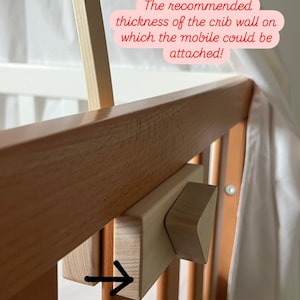 May include: Close-up of a wooden crib rail with a wooden mobile attachment. The attachment has a small, triangular piece of wood that can be adjusted to fit crib rails of different thicknesses. The text on the image reads: "The recommended thickness of the crib wall on which the mobile could be attached! Min 1,5 cm- Max 2,5 cm".