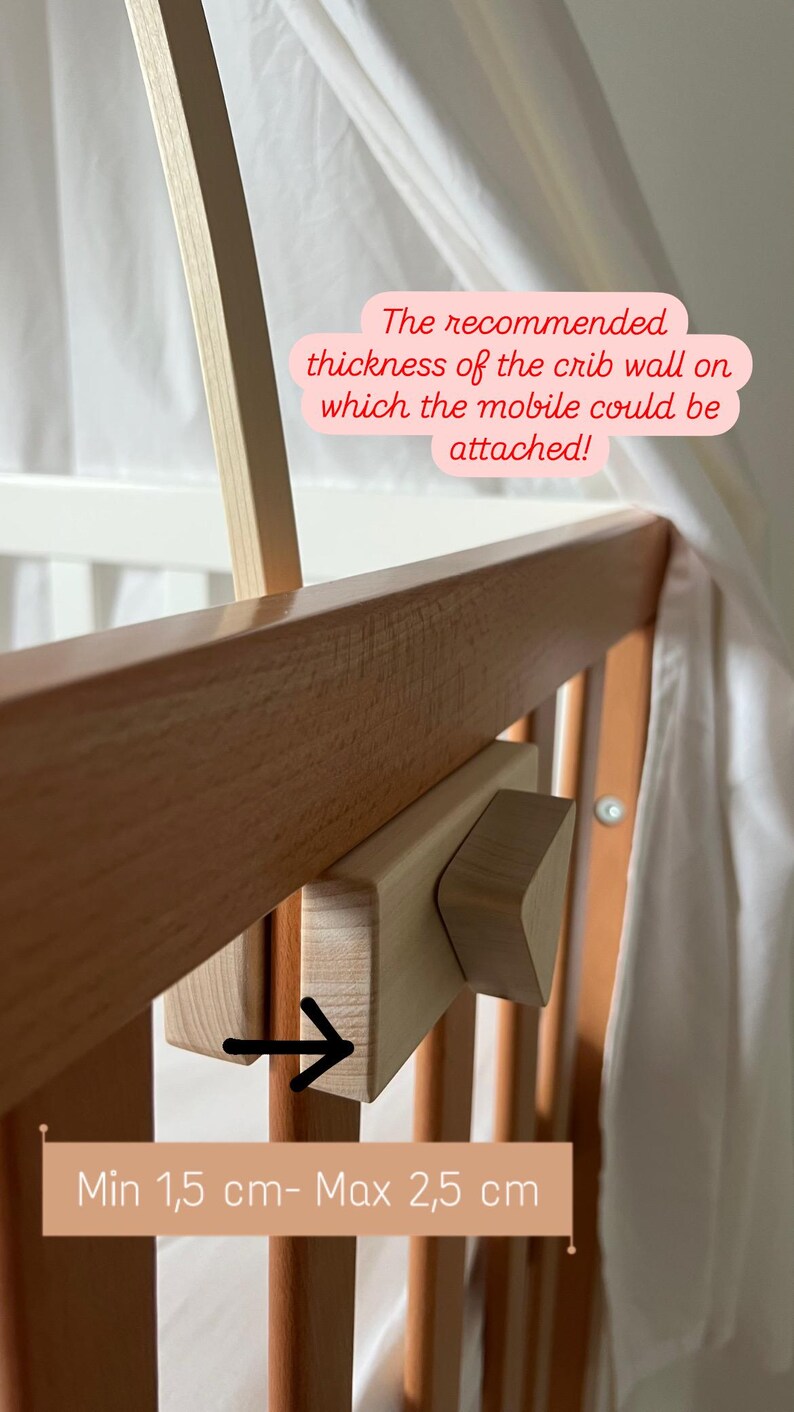 May include: Close-up of a wooden crib rail with a wooden mobile attachment. The attachment has a small, triangular piece of wood that can be adjusted to fit crib rails of different thicknesses. The text on the image reads: "The recommended thickness of the crib wall on which the mobile could be attached! Min 1,5 cm- Max 2,5 cm".