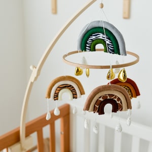 May include: A wooden baby mobile with a rainbow theme. The mobile features three rainbow-shaped fabric pieces with different animal prints, including zebra, tiger, and giraffe. The rainbows are suspended from a wooden ring and are attached to a wooden arm. The mobile also includes small white and gold teardrop-shaped decorations.