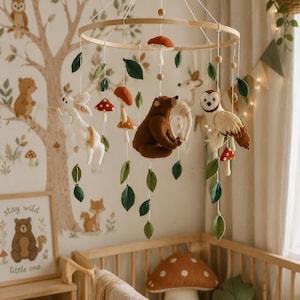 Puede incluir: Móvil para bebé de madera con animales del bosque de fieltro, incluyendo un oso, un búho y un conejo, junto con hojas verdes y setas de sombrero rojo. Cuelga sobre una cuna de madera, con un mural de pared a juego con temática de bosque.