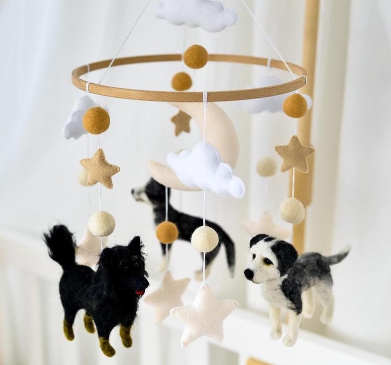 May include: A handcrafted baby mobile with felt dogs, stars, clouds, and a crescent moon. The mobile is suspended from a wooden ring by white strings. The dogs are black, white, and grey. The stars and moon are beige, and the clouds are white.
