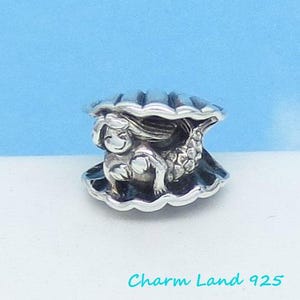 May include: Silver charm featuring a mermaid inside a seashell. The mermaid is smiling and has a tail with scales. The charm is a detailed and intricate piece of jewelry.