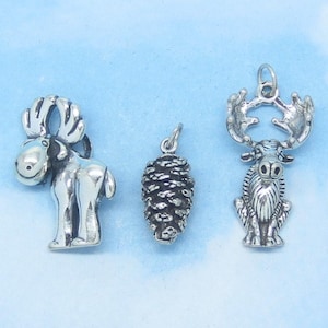 May include: Three silver-toned charms: a standing moose, a pine cone, and a seated moose. The moose charms have detailed antlers and facial features. The pine cone charm has a textured surface. All charms have a loop for attaching to a necklace or bracelet.