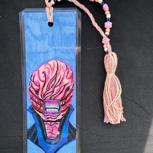 May include: A handmade bookmark featuring a monster illustration with a pink brain-like head, blue collar, and white teeth. The bookmark has a pink tassel and a beaded string with pink and blue beads.