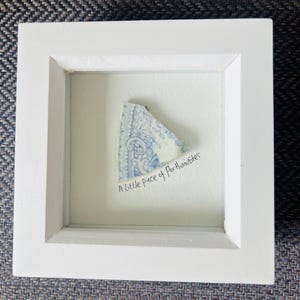 May include: A framed artwork with a white border, showcasing a fragment of blue and white patterned ceramic. The handwritten text below reads "A little piece of Porthminster".