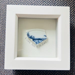 Puede incluir: Una obra de arte enmarcada en blanco con un fragmento de cerámica azul y blanca. La pieza de cerámica tiene un diseño floral y paisajístico. El texto "A little piece of St. Ives" está escrito debajo.