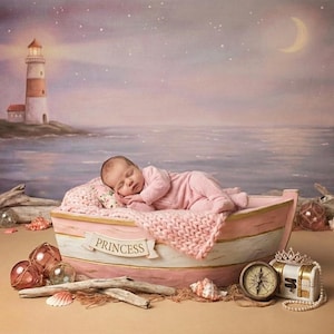 Puede incluir: Un bebé dormido con un atuendo rosa descansa en un bote rosa con la palabra "Princess". El bote está sobre una superficie arenosa con conchas marinas, una brújula y un cofre del tesoro. Un faro y una escena oceánica están en el fondo.