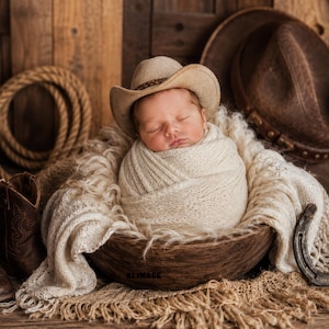 Può includere: Un neonato che dorme avvolto in una coperta di maglia color crema, con un cappello da cowboy beige, riposa all'interno di una ciotola di legno scuro. La scena include un paio di stivali di pelle marrone, un ferro di cavallo e un cappello da cowboy.