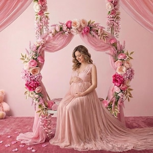 Puede incluir: Una mujer embarazada con un vestido rosa fluido está sentada en un banco, sosteniendo su vientre. Está rodeada por un arco floral y cortinas rosas, con un oso de peluche en el fondo. La escena es suave y romántica.