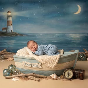 May include: A newborn baby sleeps peacefully in a small, light blue boat with the word "CAPTAIN" on the side. The nautical-themed scene includes a lighthouse, ocean backdrop, and decorative elements like shells and a compass.