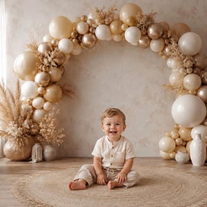 Può includere: Un bambino sorridente è seduto su un tappeto rotondo davanti a un arco di palloncini. L'arco è composto da palloncini bianchi, dorati e beige. Erba della pampa secca e vasi decorativi sono ai lati dell'arco. Il bambino indossa una camicia e pantaloni color crema.
