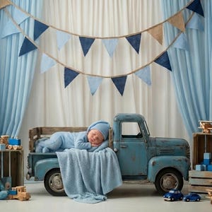 May include: A baby in a blue outfit and hat is sleeping in the back of a vintage blue truck. The scene is decorated with blue and tan triangle banners, wooden toy blocks, and a teddy bear. The truck is a charming prop for a newborn photoshoot.