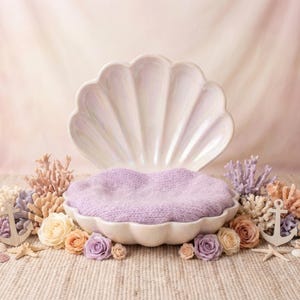 May include: A seashell-shaped prop for newborn photography, with a soft lavender nest. The scene is decorated with coral, flowers, anchors, and seashells, creating a dreamy, underwater theme.